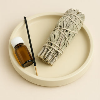 A digital photograph displays a smudging stick, an incense stick, and a small amber glass bottle arranged on a round cream-colored tray against a neutral beige background, with soft lighting highlighting their textures.